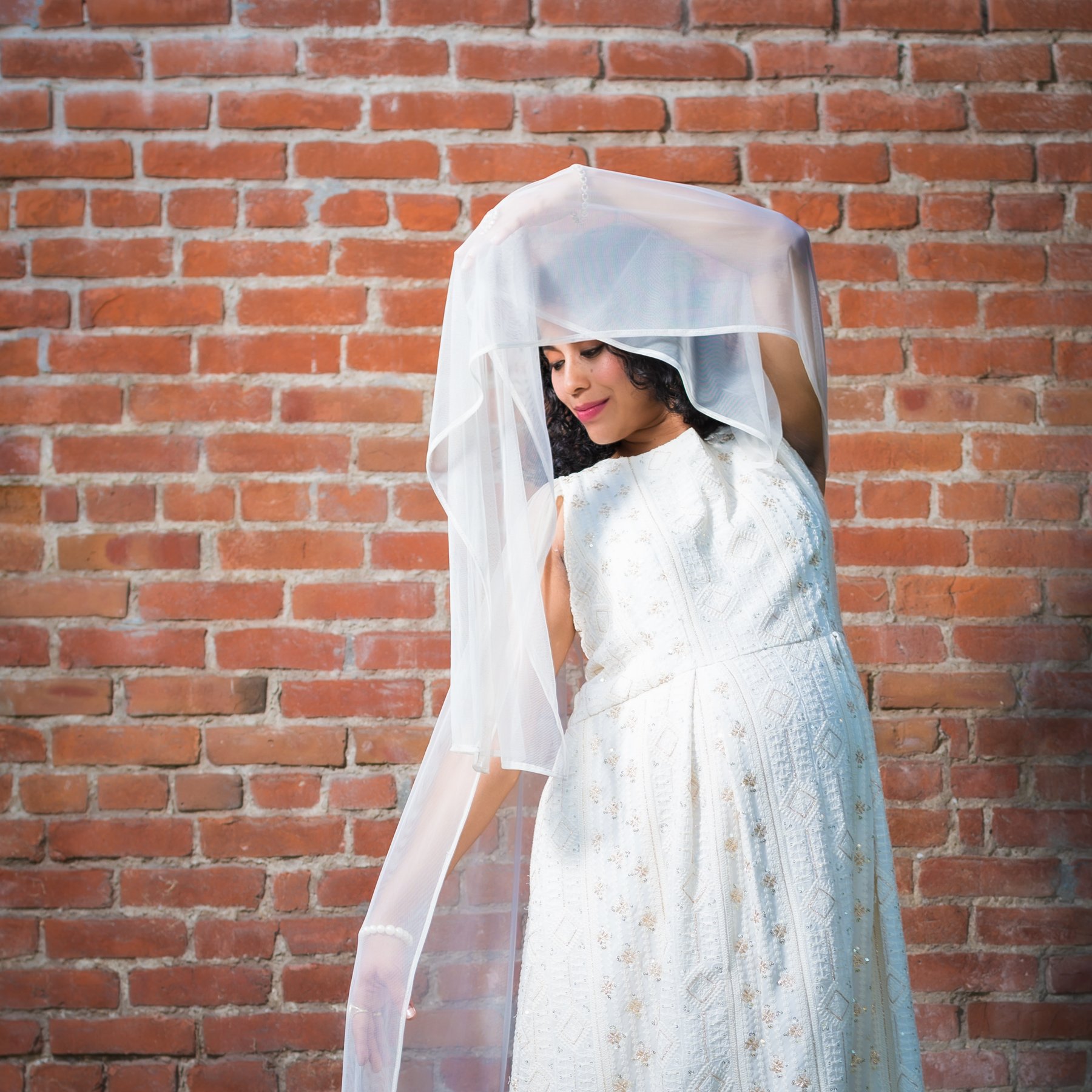 Different setup entirely. Ivory chikankari against red brick, veil lifted and caught mid air, and she's looking down and away with a small smile. The color contrast is doing most of the work here... warm brick, cool white tulle, nothing in between.